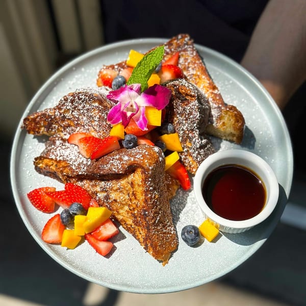 French toast with powdered sugar, strawberries, blueberries, mango, mint, and an orchid, with syrup.