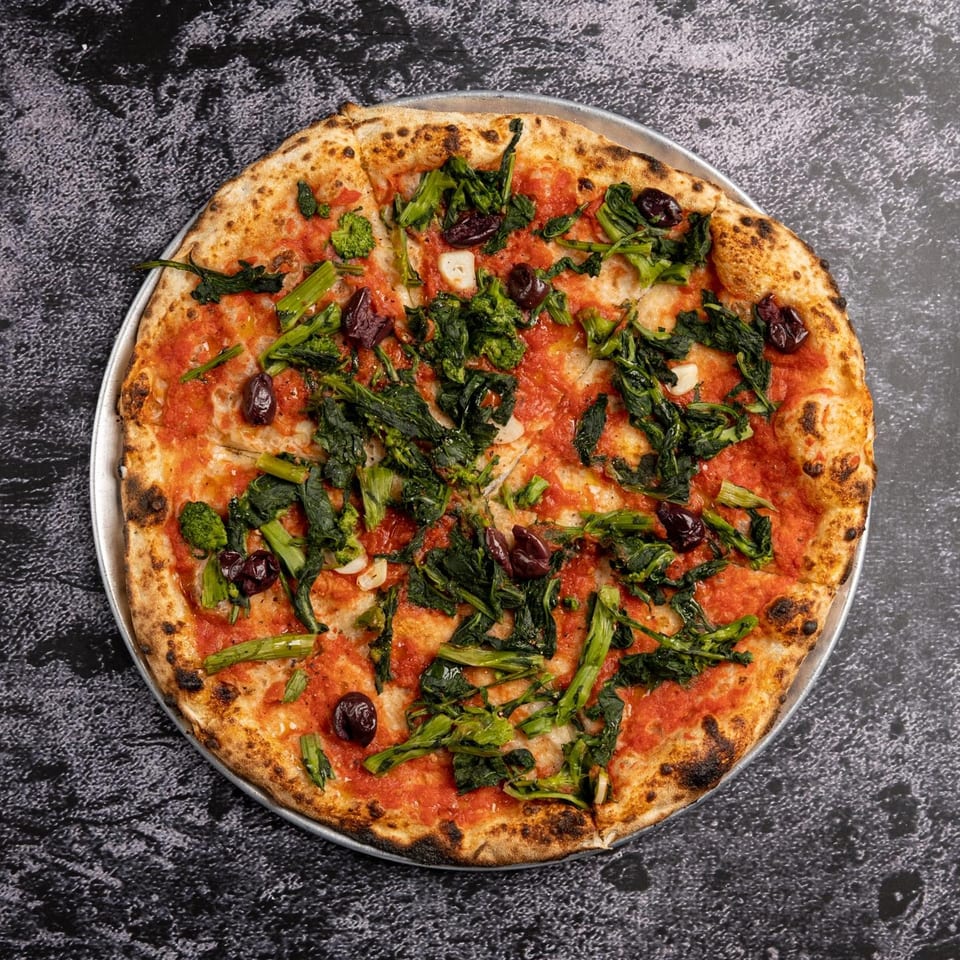 Black Oak on Fifth's Italian pizza with tomato sauce, green vegetables, and olives on a dark surface.