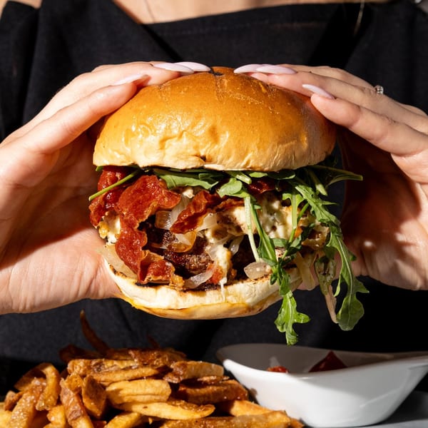 Hands holding a burger with bacon, onions, and arugula, with fries and dipping sauce below.