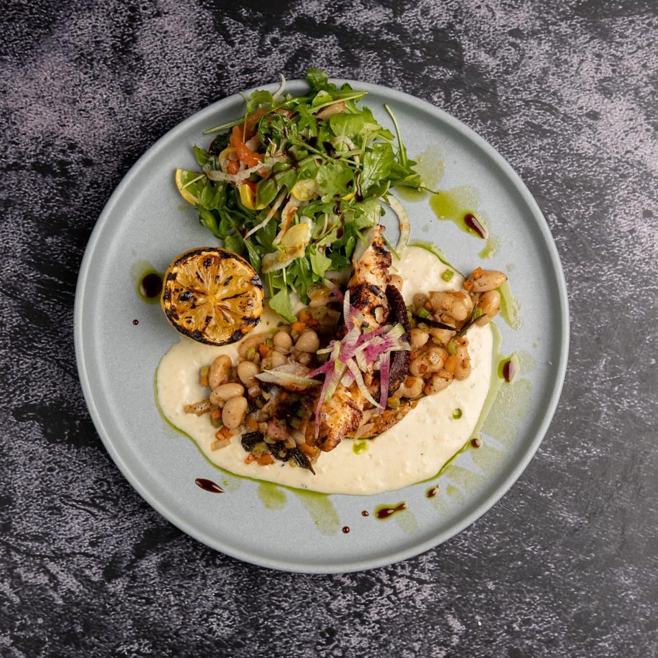Grilled octopus with white beans, arugula salad, and a charred lemon on a light blue plate.